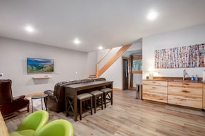 Ein modernes Wohnzimmer mit einer Couch, einem Esstisch mit Hockern, einem an der Wand befestigten Fernseher und einer Küchenzeile mit Holzschränken und einer Spüle.
