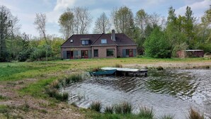 Una casa de dos pisos con techo inclinado, una chimenea y un pequeño estanque con un muelle.