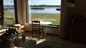 Una habitación con un amplio ventanal con vistas a un lago, una silla de madera, una mesa y un pequeño carro con leña.