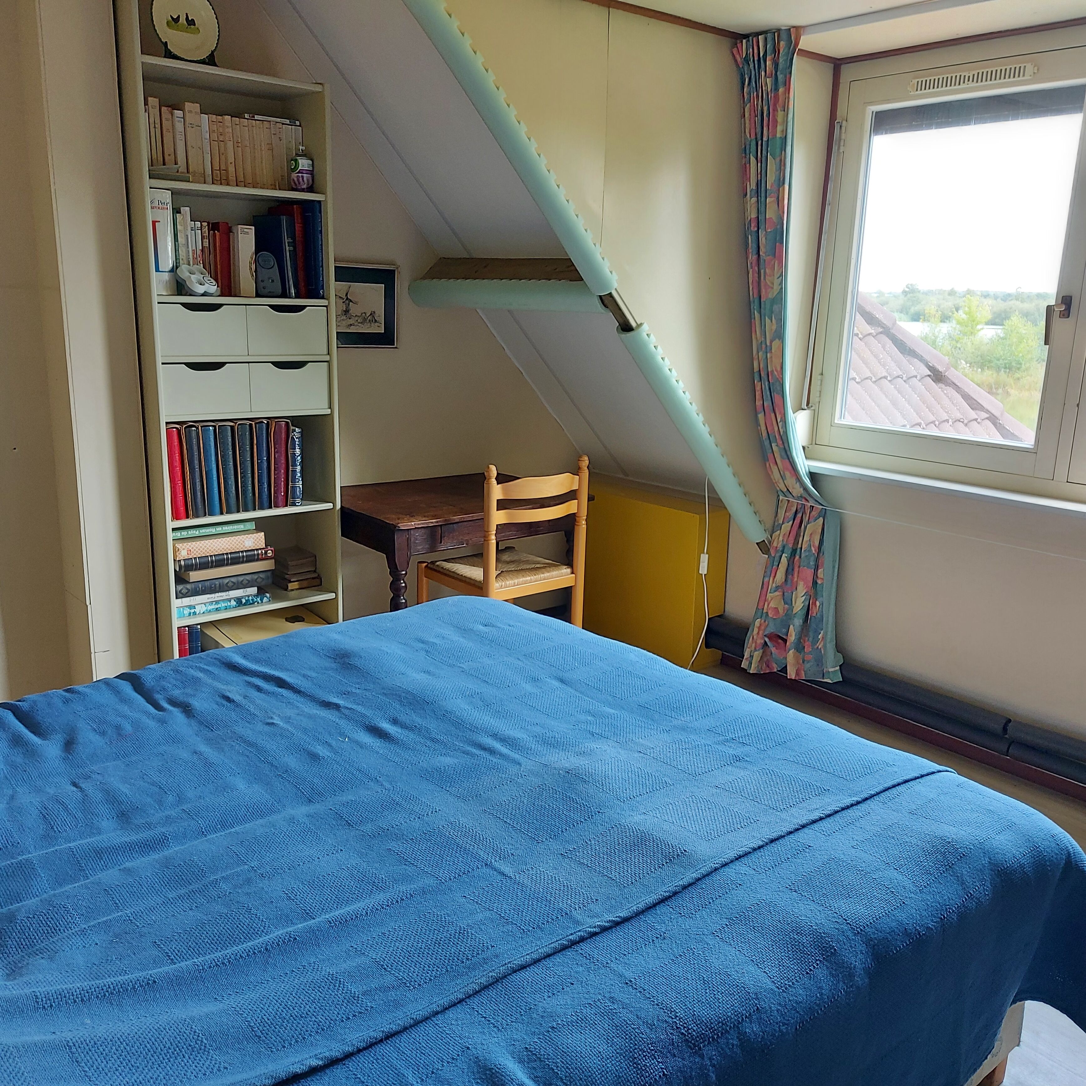Een slaapkamer met een bed, een houten stoel, een boekenkast en een raam met gordijnen.