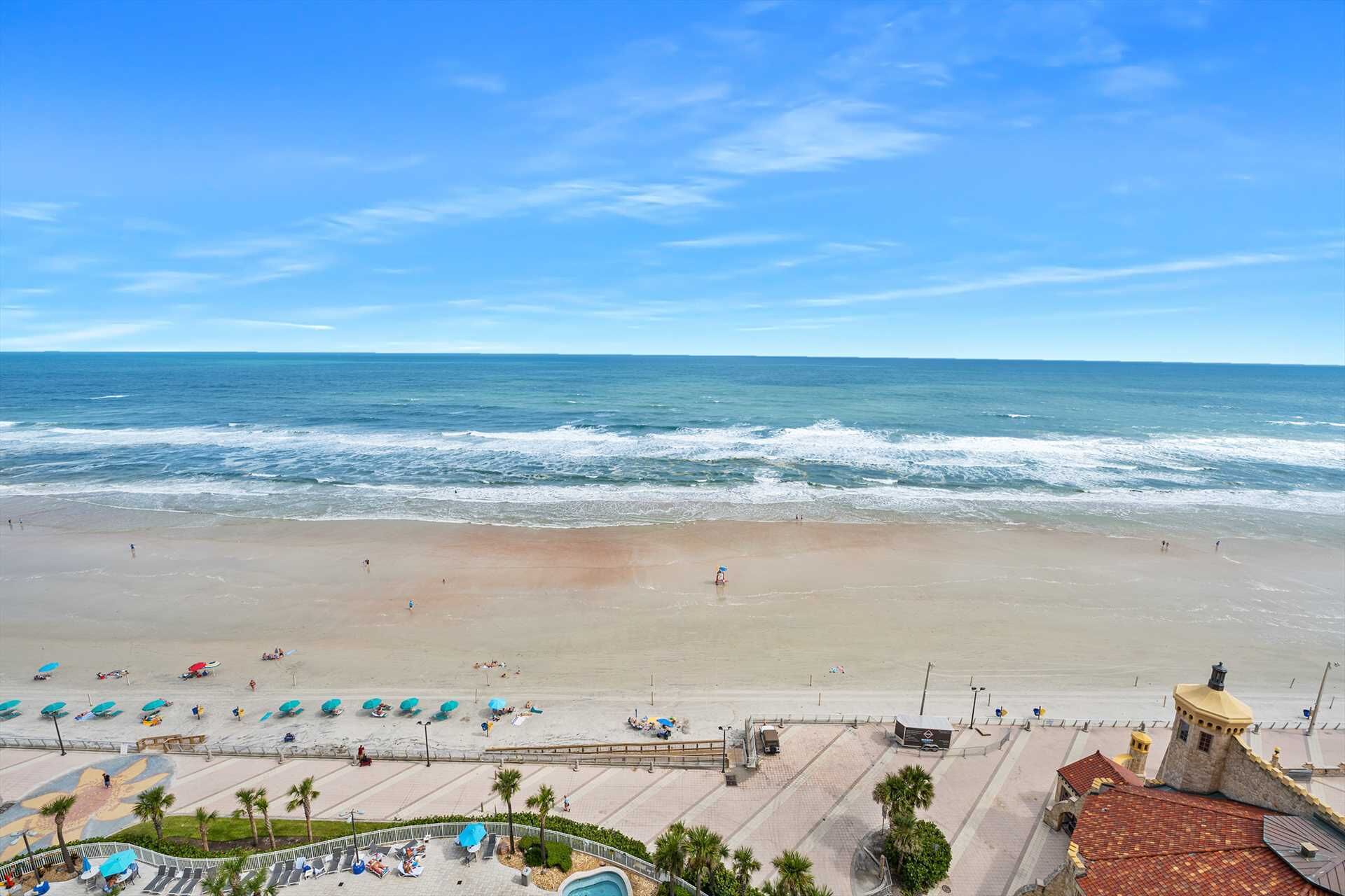 A beach with a clear sky, a sandy shore, and a few people. There are umbrellas and lounge chairs along the beach.