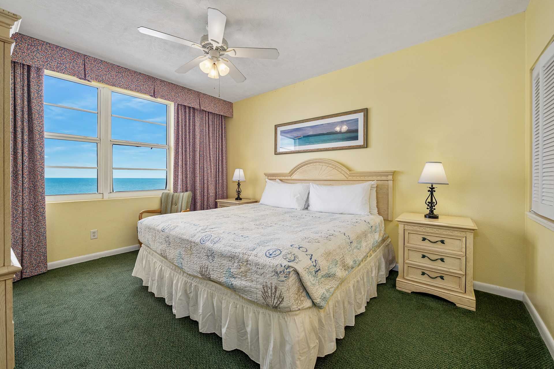 A bedroom with a bed, two bedside tables with lamps, a ceiling fan, a window with curtains, and a painting on the wall.