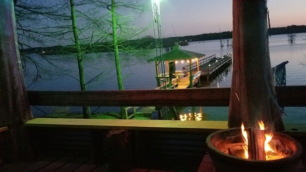 Una terraza de madera con un brasero, con vista a un muelle y un pequeño pabellón junto al agua.