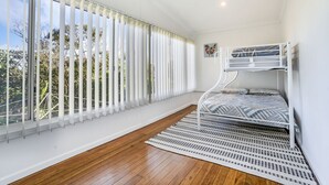 Una habitación con literas, ventanas grandes con persianas, piso de madera y pared blanca.