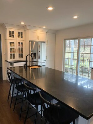 A modern kitchen with a large island, white cabinetry, stainless steel appliances, and a view through large windows.