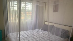 Ett sovrum med en säng som är täckt av ett myggnät, ett fönster med gardiner och en liten hylla som är monterad på väggen.