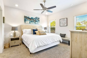 A bedroom with a bed, bedside tables, a ceiling fan, artwork, a window, and a bench.