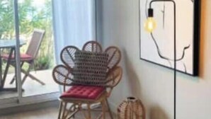 Een kamer met uitzicht op groen, een rieten stoel, een vloerlamp en een abstract kunstwerk aan de muur.
