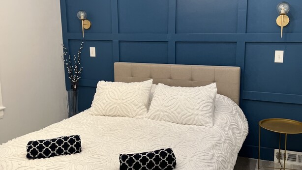 Une chambre à coucher avec un grand lit, des murs recouverts de lambris bleus, un lustre moderne et une petite table d’appoint.