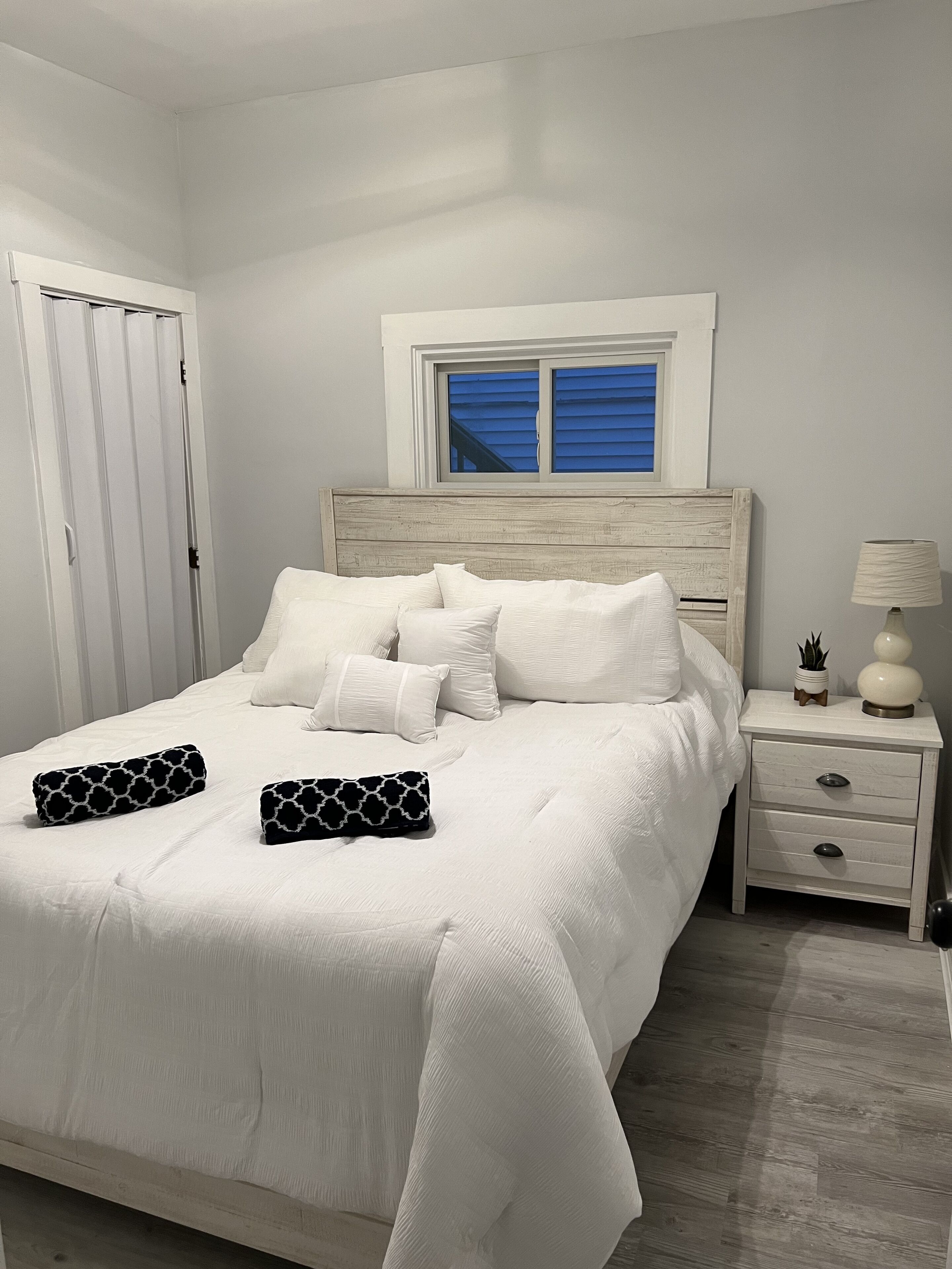 Un lit bien fait, avec une literie blanche et deux oreillers noirs ornés d’un motif blanc. On y voit une tête de lit en bois, une petite table de chevet avec une lampe, et une fenêtre avec des volets bleus.