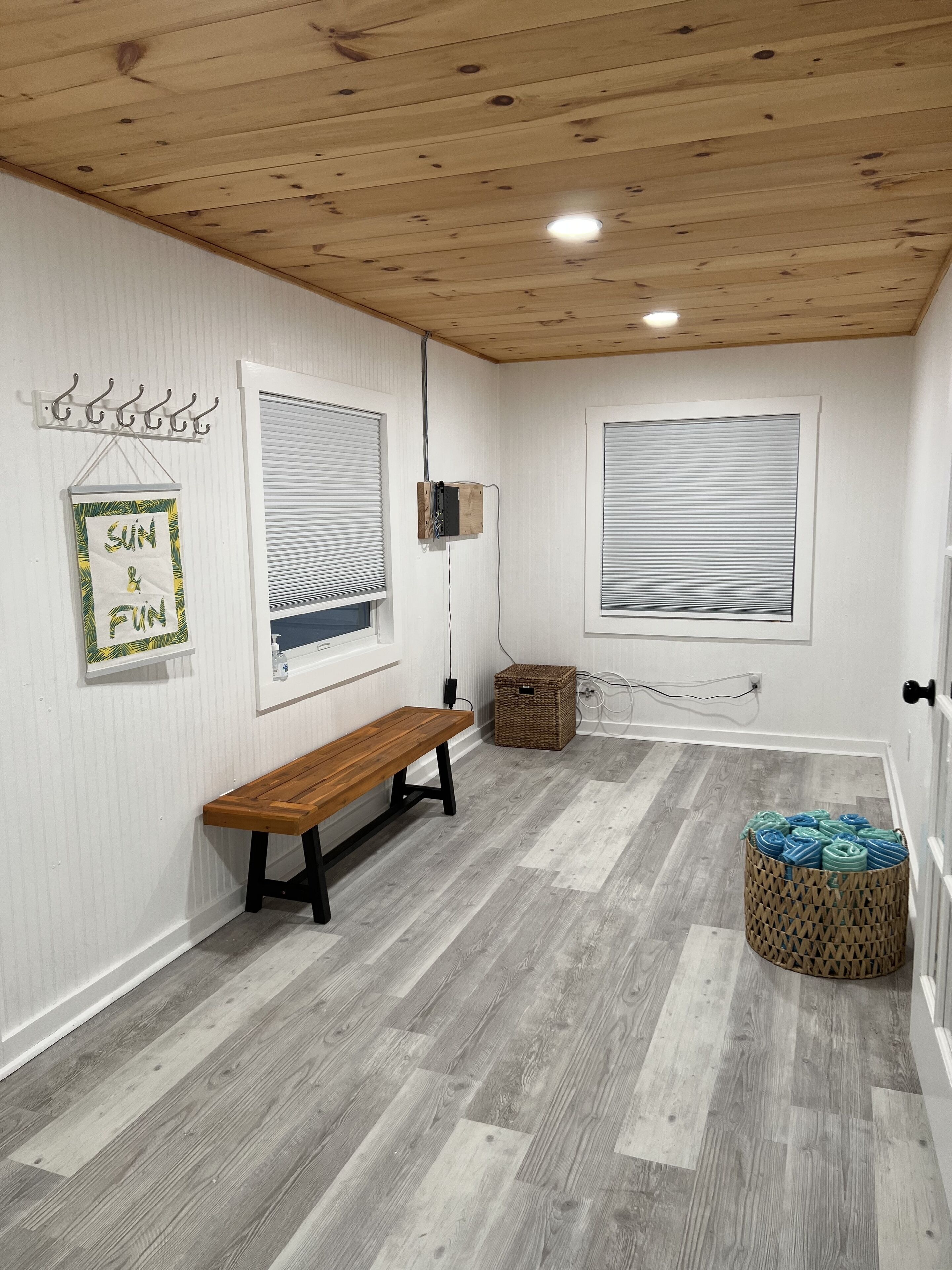 A room with a wooden ceiling, white walls, a bench, a wicker basket, and a wall-mounted shelf with a sign.