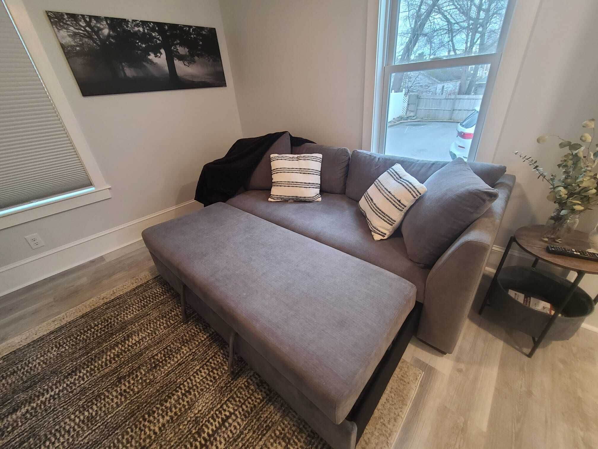 En moderne stue med en grå sofa, to stribede puder, et sort pled og et lille sofabord med en vase og en fjernbetjening.