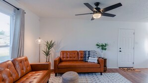 A living room with a brown leather sofa, a round ottoman, a patterned rug, a ceiling fan, a floor lamp, and a door with a doormat. - Instant Book, Clean & Cozy - minutes to Kalahari (Round Rock)