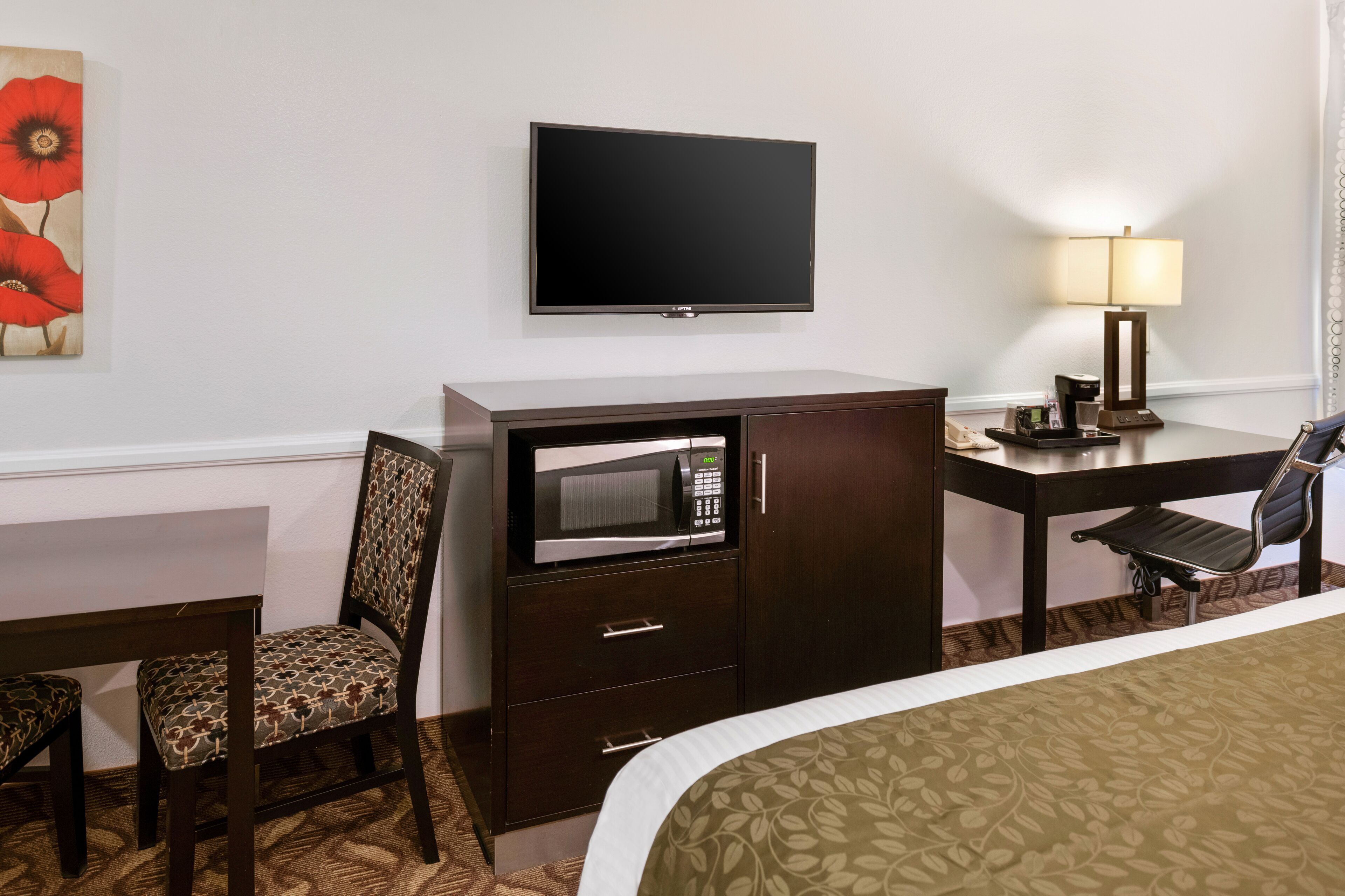 A hotel room with a bed, a desk with a lamp, a microwave, a TV, and a chair.