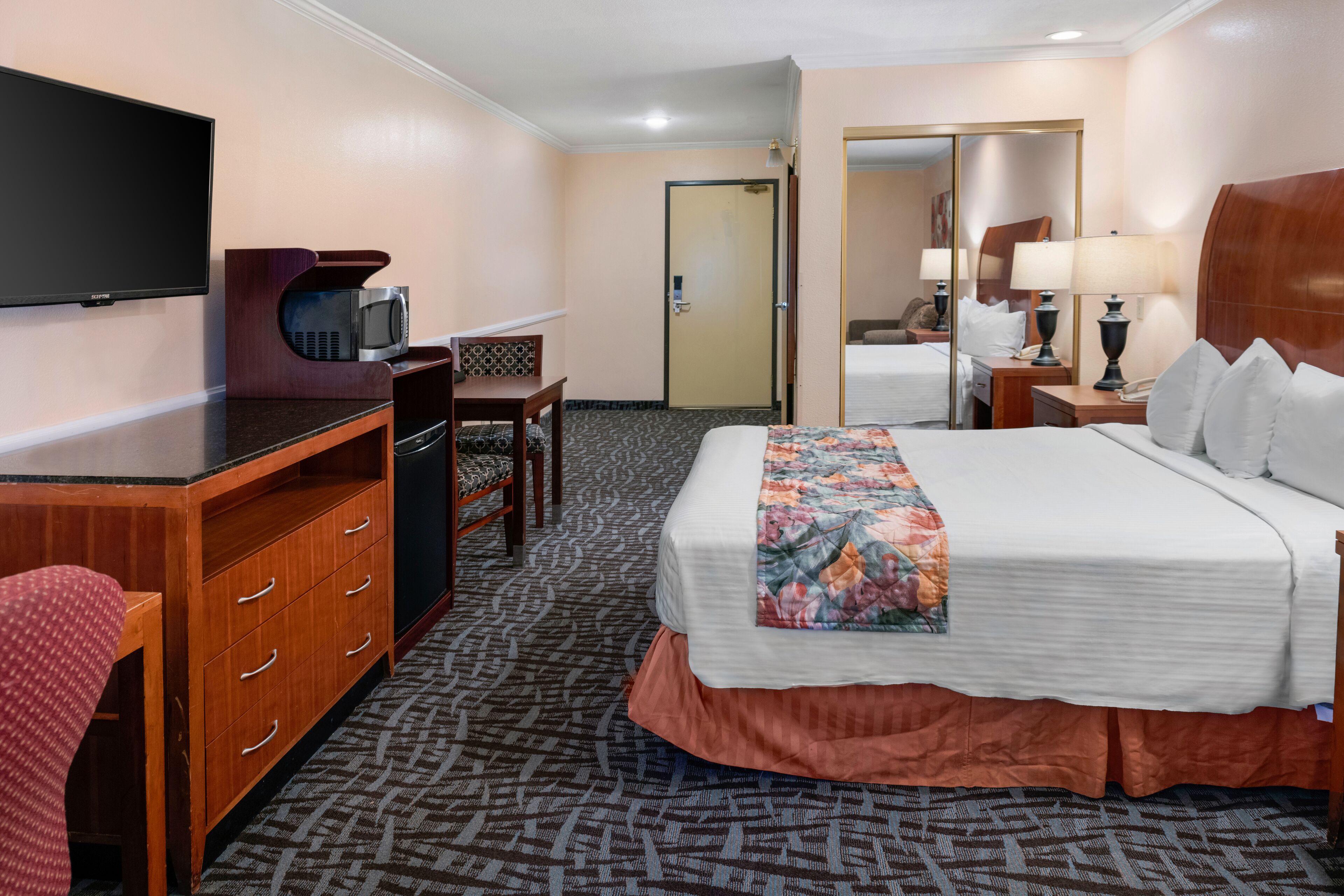 A hotel room with a bed, a desk with a microwave, a TV, a mirror, and a door.