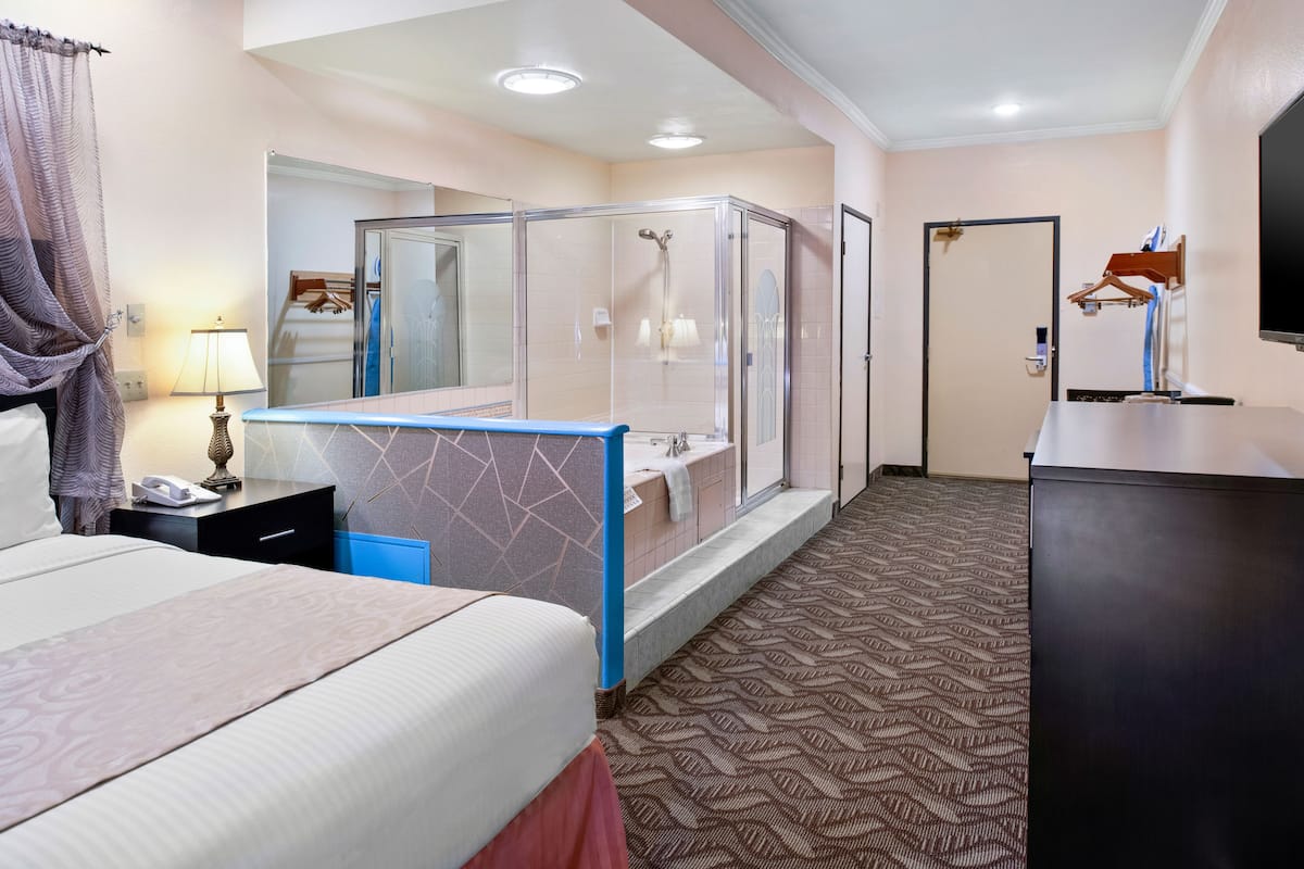 A hotel room with a bed, a bedside table with a lamp, a shower area, and a TV on a stand.