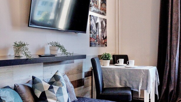 Una sala moderna con un televisor de pantalla plana grande montado en la pared, un sofá con cojines estampados, una mesa de comedor con sillas y una planta en maceta.