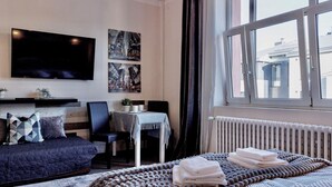 Una habitación de hotel moderna con una cama grande, un sofá, un televisor de pantalla plana montado en la pared, una mesa de comedor con sillas y una ventana con vistas a edificios.