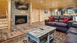 Interior acogedor de una cabaña de troncos con chimenea de piedra, pisos de madera, un sofá de cuero y un ventilador de techo.