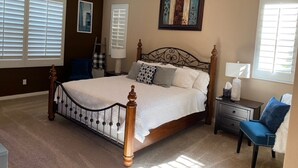 Une chambre à coucher avec un grand lit, un ventilateur de plafond, une table de chevet, une chaise et une fenêtre avec des stores.