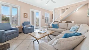 A spacious living room with a beige sofa, blue armchairs, and a wooden coffee table. A ceiling fan and a staircase with a railing are visible. - Ocean Terrace-Large beach house with private pool (Gulf Shores)