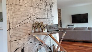 A living room with a large map mural on the wall, a glass coffee table with a sign reading "ILLINOIS," a gray sofa, and a flat-screen TV. - Oakton St. Inn, Evanston near Northwestern, Loyola and Chicago (Evanston)