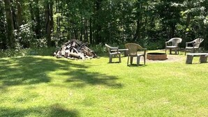 Un espacio al aire libre con césped, una pila de leña, varias sillas de plástico y una mesa redonda.