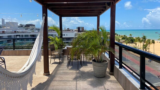 A balcony with a hammock, potted plants, and a view of the beach and cityscape. - Bela Hospedagem - Marinas Cabo Branco (João Pessoa)