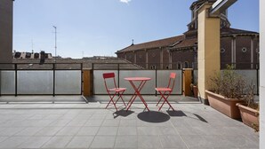 Una terraza con sillas rojas y una mesa, un edificio de ladrillo con una cruz al fondo, y un cielo despejado.