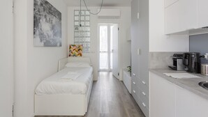 Un dormitorio con cama, un cojín de colores, un cuadro colgado en la pared y una zona de cocina con cafetera.