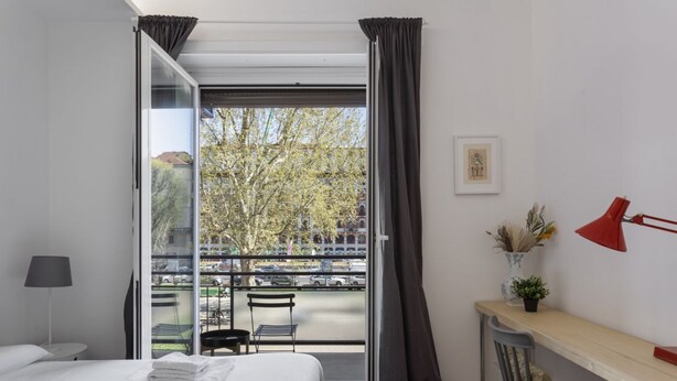 Ein Hotelzimmer mit Bett, Schreibtisch, Stuhl, Lampe und Blick ins Freie.