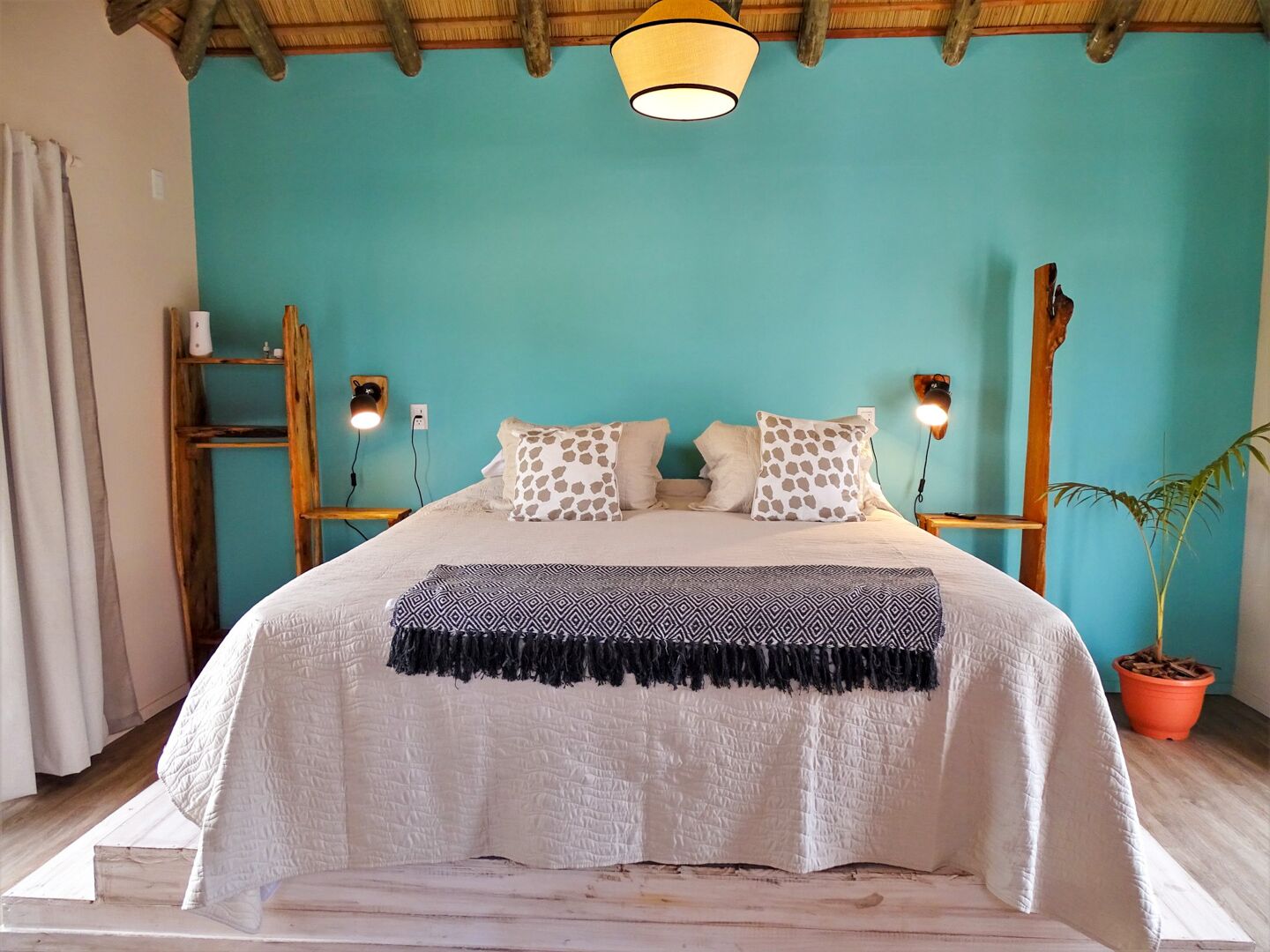 A bed with a white quilt, patterned pillows, and a black fringe throw. There are two wall-mounted lamps on either side of the bed.