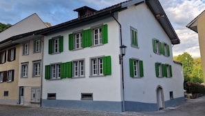 A multi-story building with green shutters, a cobblestone street, and a no parking sign. - "VILLA HI" apartment parking space included  (Weesen)