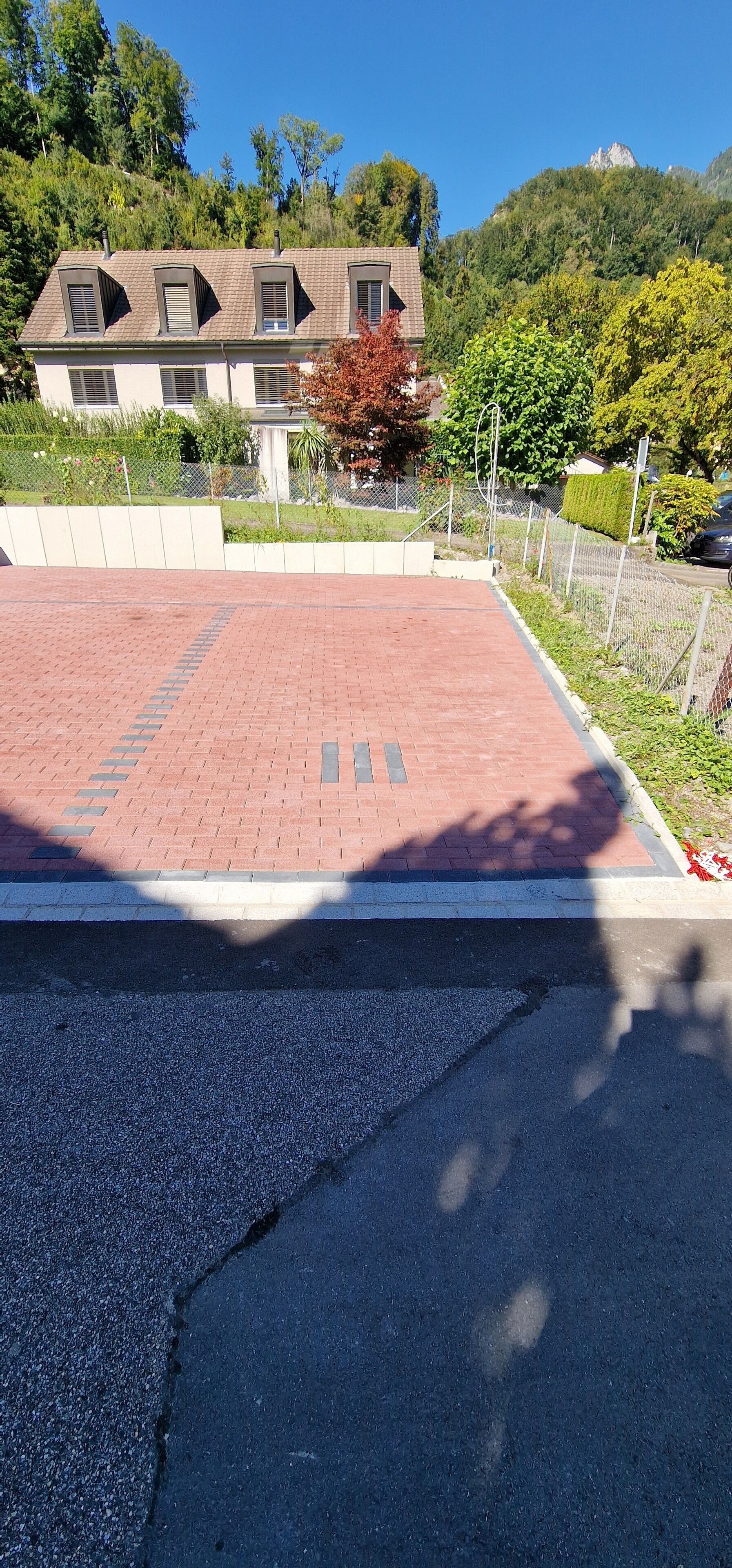 Ein gepflasterter Bereich mit Ziegelmuster, eine Straße und ein Gebäude mit vielen Fenstern und einem geneigten Dach.