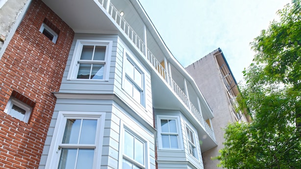 Um edifício angular com fachada de tijolo, várias janelas e uma varanda. Há mesas e cadeiras de madeira dispostas no chão.