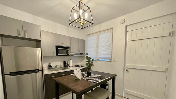 A compact kitchen with a stainless steel refrigerator, a microwave, and a sink. There's a dining table with a placemat and a potted plant.