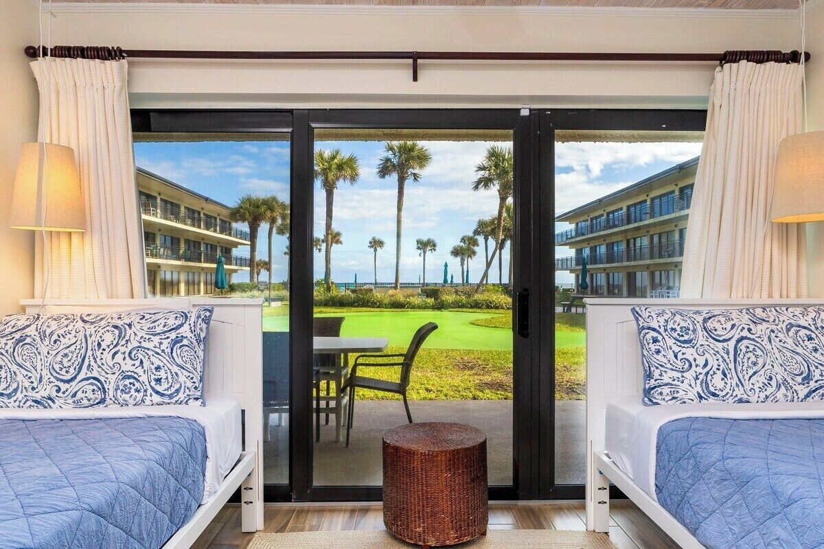 A hotel room with a large sliding glass door leading to a golf course view, two beds with patterned pillows, a wicker ottoman, and a table with chairs.