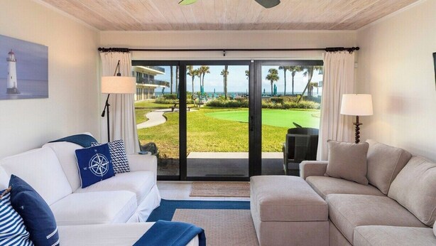 Una habitación con ventilador de techo, dos sofás, un ventanal con vista a un campo de golf y un cuadro de un faro.