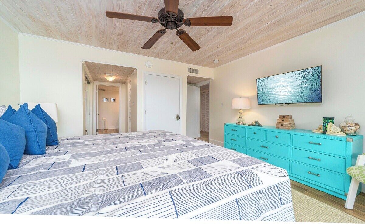 Een slaapkamer met een bed, een plafondventilator, een televisie, een turquoise dressoir en een deur naar een andere kamer.