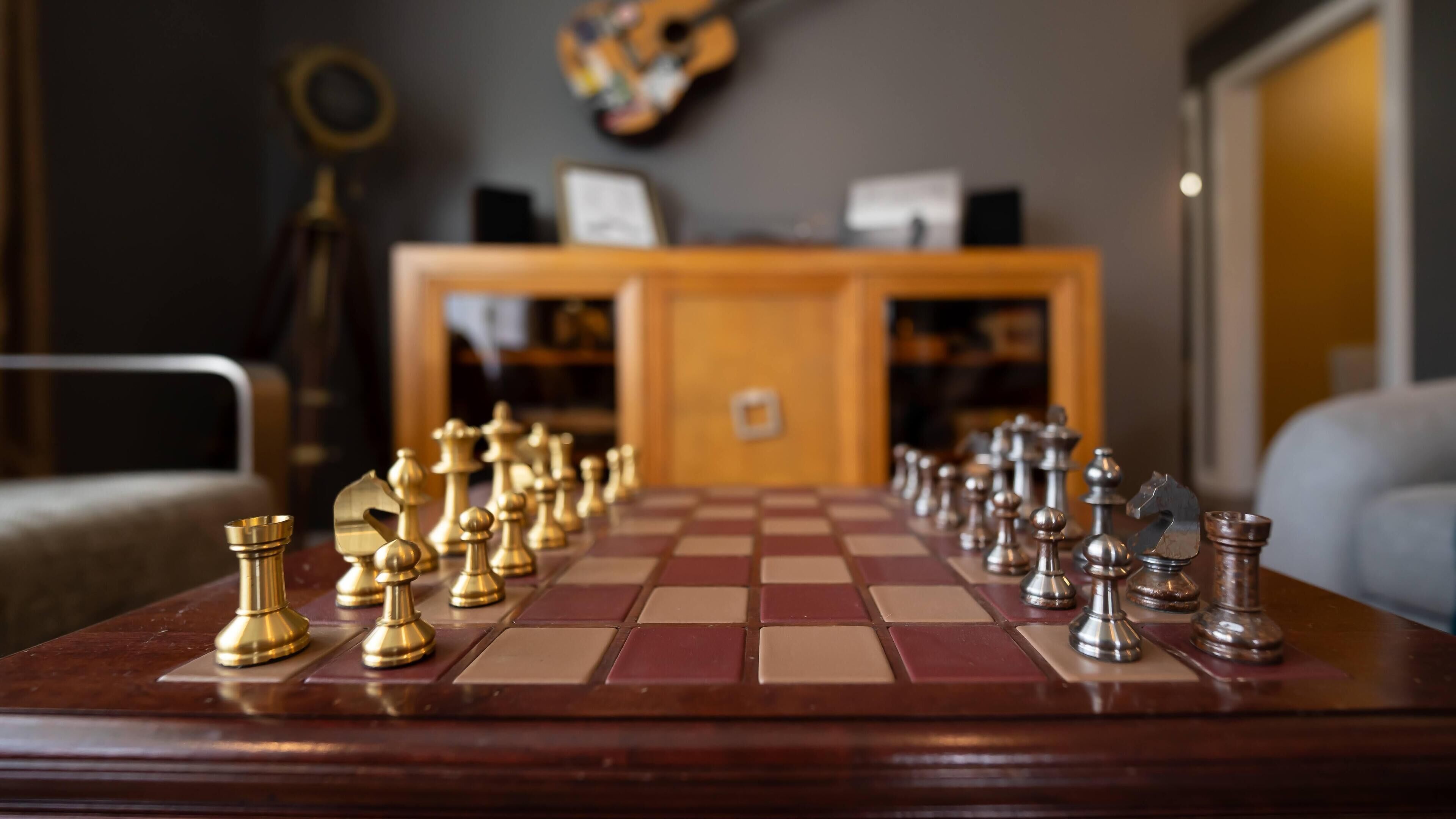Een schaakbord met stukken in een kamer, met een houten kast en een gitaar op de achtergrond.