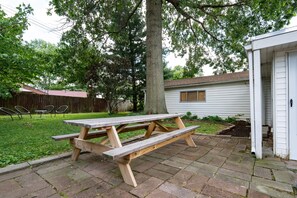 Una mesa de picnic de madera en un patio trasero con un adoquín, una casa blanca y una cerca de madera al fondo.