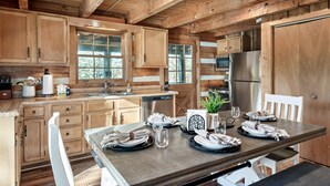A wooden cabin kitchen with a dining table set for four, wooden cabinets, a refrigerator, and a window providing natural light.