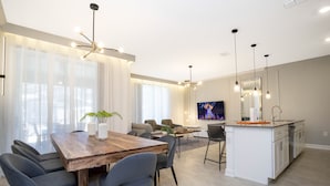A modern kitchen with a central island, a dining area with a wooden table and chairs, and a living area with sofas and a TV.