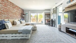 Una sala de estar moderna con una pared de ladrillo, un conjunto de sofás, un televisor y un balcón con mobiliario exterior.