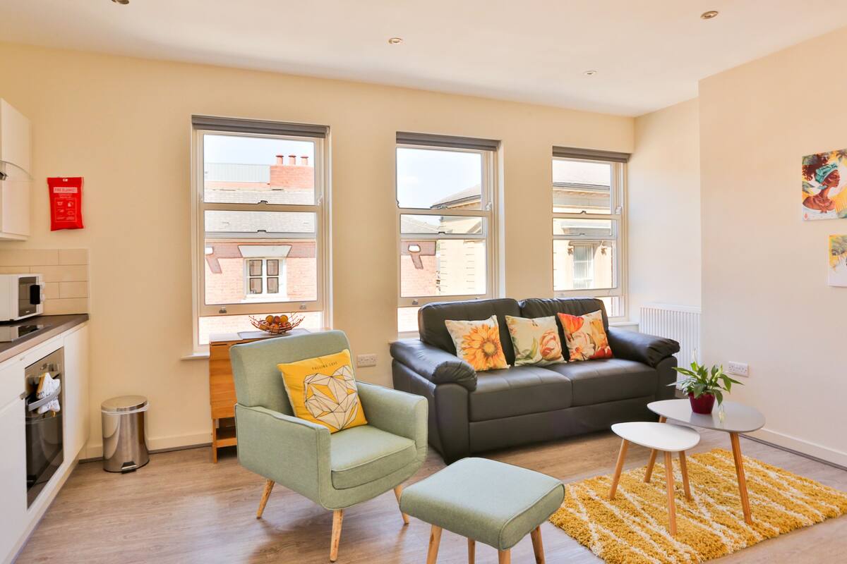 A modern living room with a sofa, armchair, and ottoman, a small table with a plant, and large windows offering views of the outside.