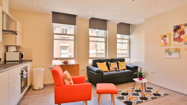 Un apartamento moderno con cocina, sala de estar con sofá y una silla naranja brillante, ventanas amplias y obras de arte en las paredes.