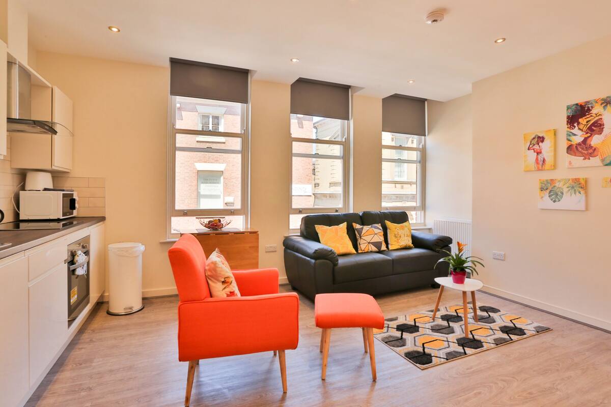 A modern apartment with a kitchen, a living area with a sofa and a bright orange chair, large windows, and artwork on the walls.