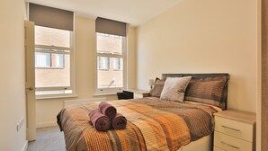Un dormitorio con cama, dos ventanas, un escritorio y una mesita de noche.