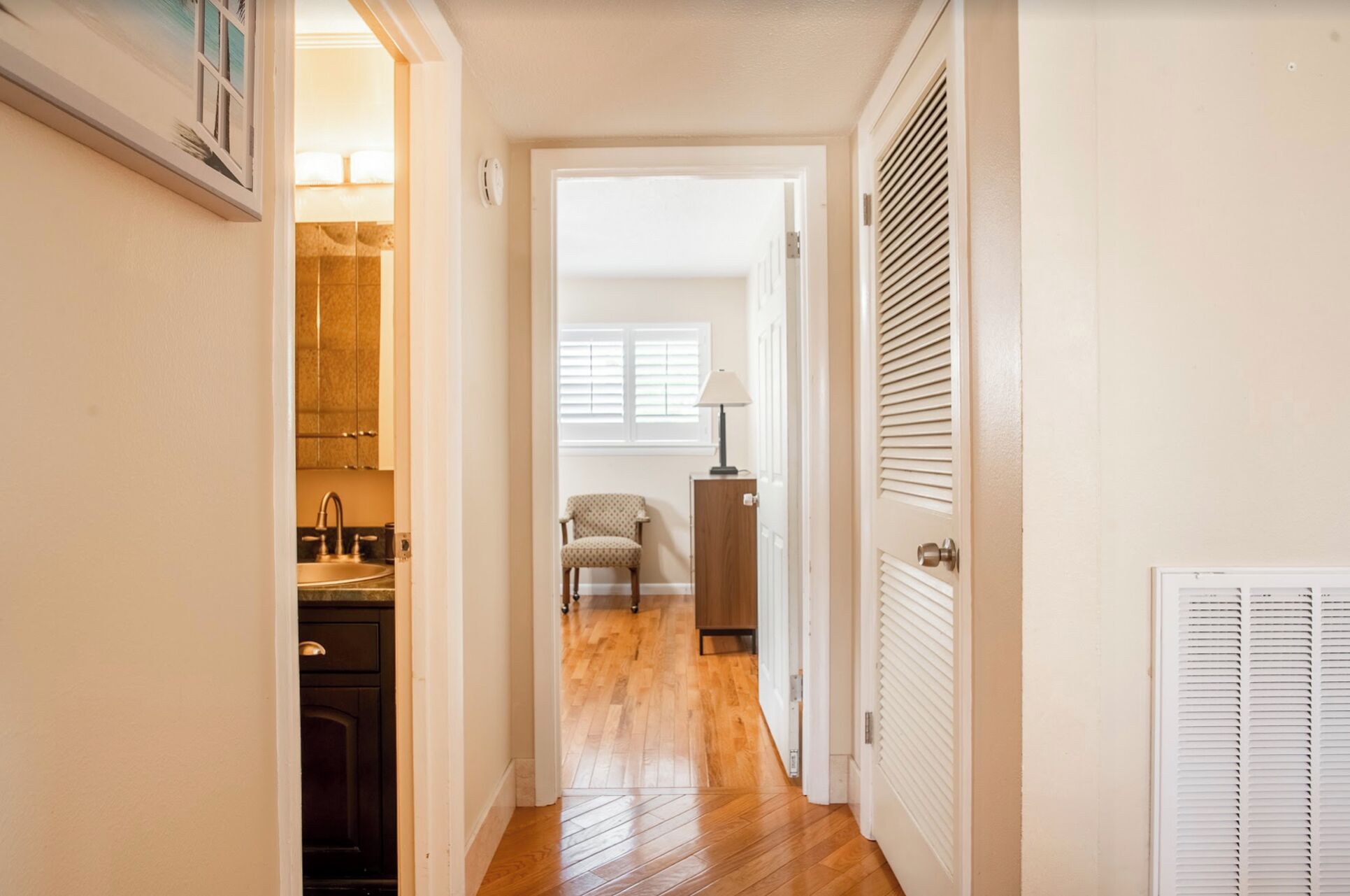 A hallway with a wooden floor, a chair, a lamp, and a door with louvered shutters.