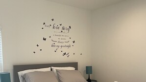 A neatly made bed with a blue and white duvet, a decorative wall with a love story quote, and a small bedside table with a lamp.